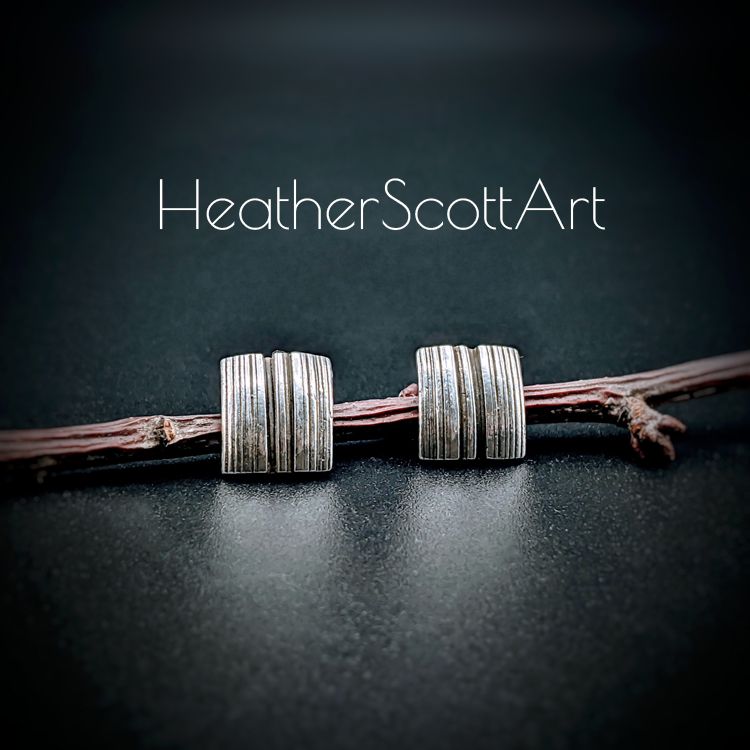 A pair of rectangle stud earrings with a vertical linear texture design in black and silver. There is a twig prop behind them and the words Heather Scott Art above them.