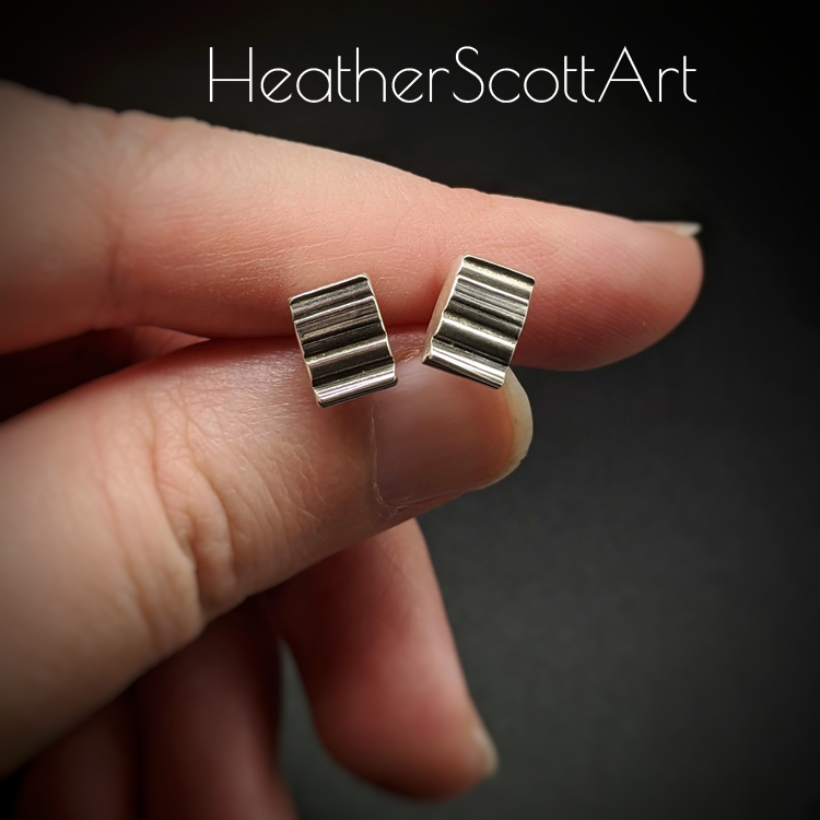 A pair of small rectangle stud earrings with a horizontal linear texture design in black and silver are being held between the artists fingers. The hand sits on a black background with the words Heather Scott Art above it.