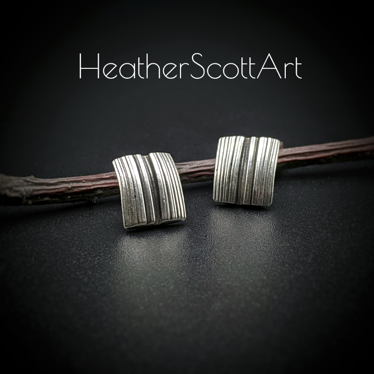 A pair of rectangle stud earrings with a vertical linear texture design in black and silver. There is a twig prop behind them and the words Heather Scott Art above them.