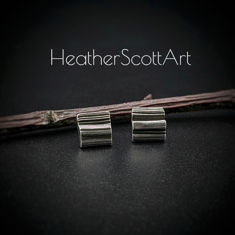 A pair of small rectangle stud earrings with a linear texture design in black and silver. They sit in front of a textured twig on a black background with the words Heather Scott Art above them.