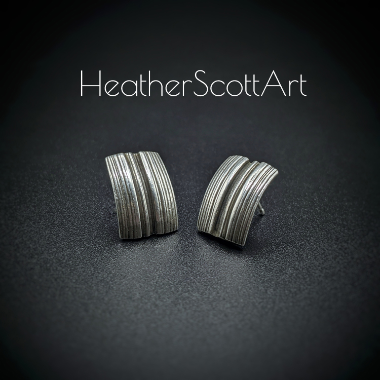A pair of rectangle stud earrings sitting at a slight angle toward one another with a vertical linear texture design in black and silver. The rectangles are slightly curved. They sit on a black background with the words Heather Scott Art above them.